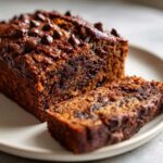 Close-up of a moist Vegan Chocolate Banana Bread loaf with a slice cut, topped with melted chocolate chips.