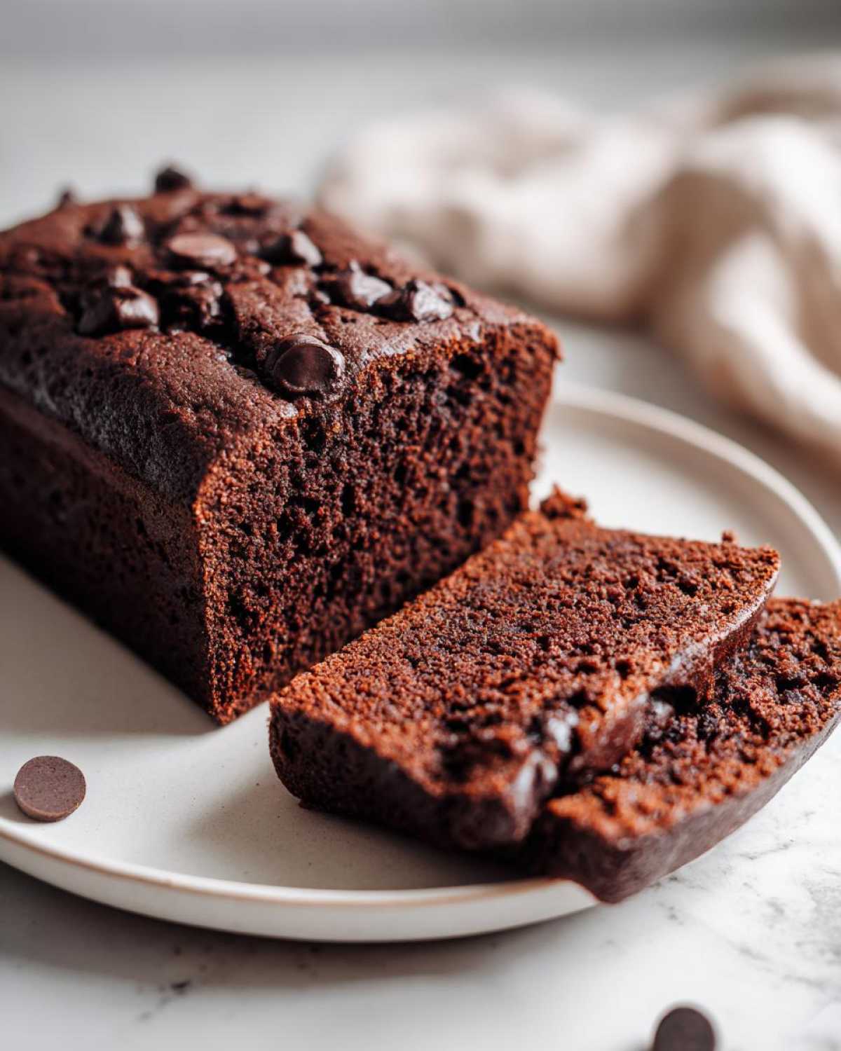 A loaf of moist Vegan Chocolate Banana Bread, partially sliced, topped with chocolate chips on a white plate.