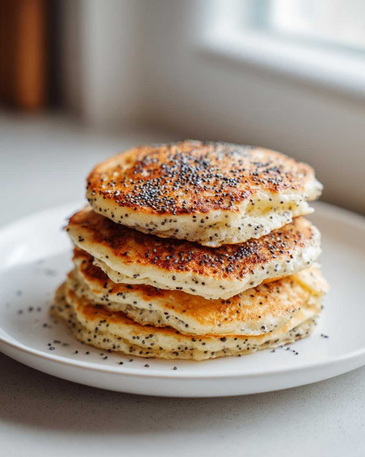A tall stack of four fluffy Vegan Lemon Poppy Seed Pancakes topped generously with poppy seeds on a white plate.