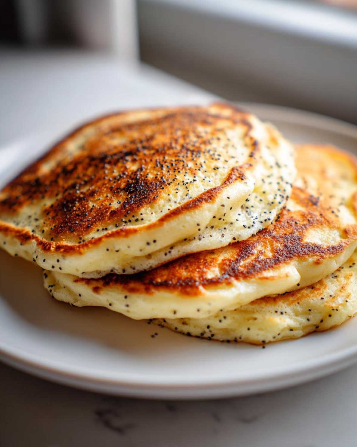 A stack of three fluffy Vegan Lemon Poppy Seed Pancakes topped with visible poppy seeds.
