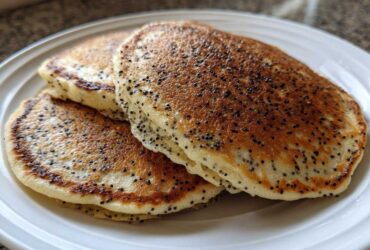 A stack of three fluffy Vegan Lemon Poppy Seed Pancakes with visible poppy seeds on a white plate.