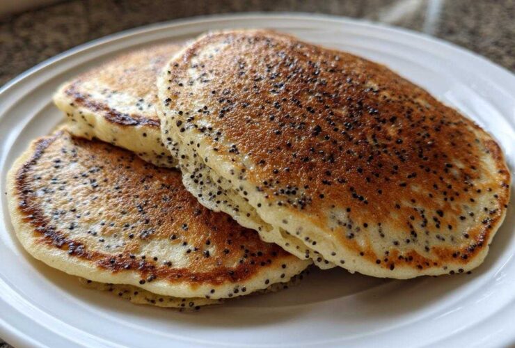 A stack of three fluffy Vegan Lemon Poppy Seed Pancakes with visible poppy seeds on a white plate.