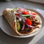 A delicious Vegan Mushroom Shawarma wrap filled with shredded mushrooms, cucumber, tomato, red onion, and drizzled with white sauce, served on a plate.