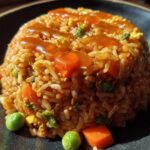 A molded serving of Vegetable Fried Rice with Sesame Ginger Sauce, featuring carrots and peas, on a dark plate.