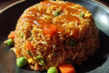 A molded serving of Vegetable Fried Rice with Sesame Ginger Sauce, featuring carrots and peas, on a dark plate.