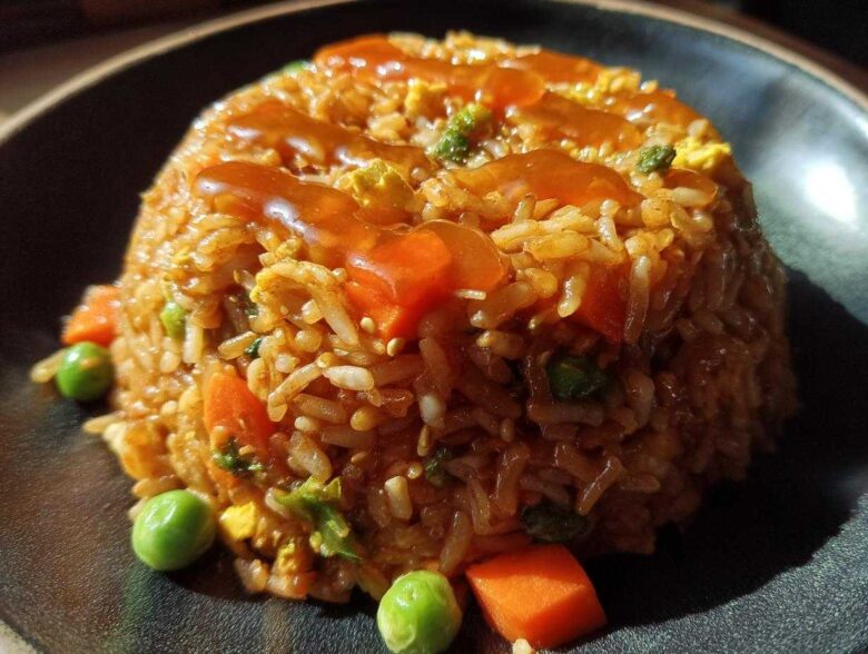A molded serving of Vegetable Fried Rice with Sesame Ginger Sauce, featuring carrots and peas, on a dark plate.