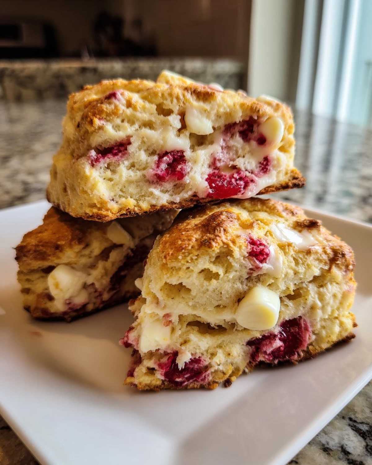 Close-up of three flaky White Chocolate Raspberry Scones stacked on a white plate, showing melted white chocolate chips and bright red raspberries inside.