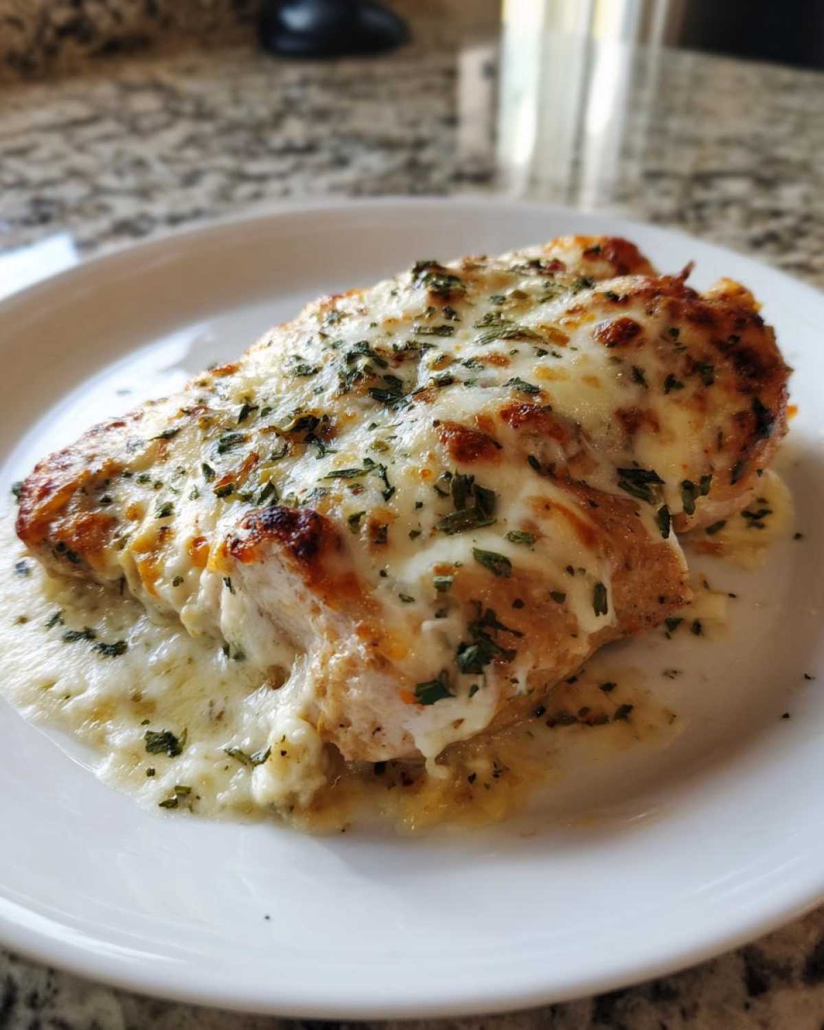 A close-up of Cheesy Baked Chicken with Cream Sauce, topped with melted cheese and herbs, served on a white plate.