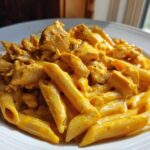 A close-up shot of penne pasta mixed with chunks of chicken in a rich, orange-hued Cheesy Chicken Pasta with Creamy Sauce, served on a white plate.