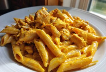 A close-up shot of penne pasta mixed with chunks of chicken in a rich, orange-hued Cheesy Chicken Pasta with Creamy Sauce, served on a white plate.
