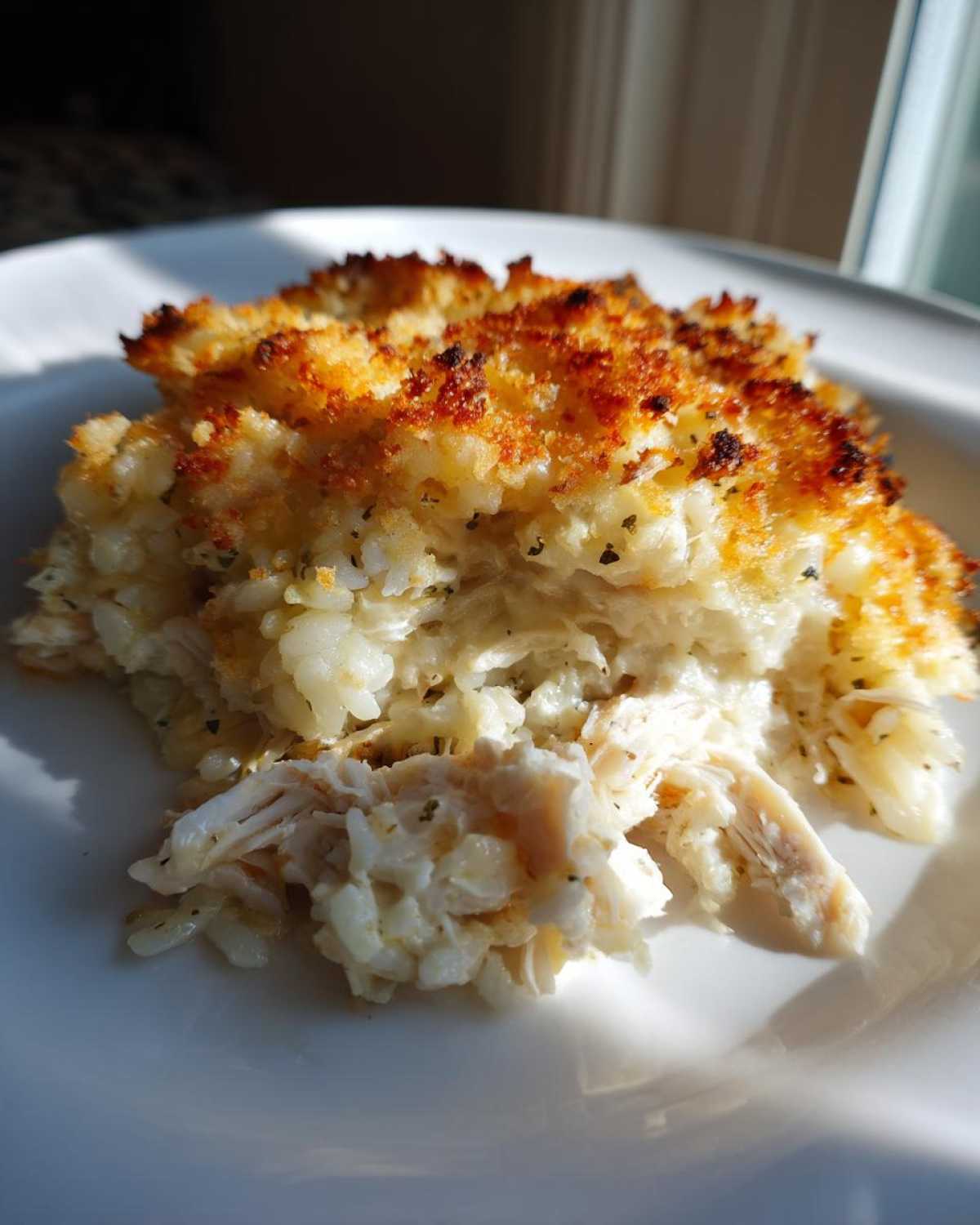 A close-up serving of Cheesy Chicken and Rice Bake with Golden Top showing shredded chicken and creamy rice beneath a crispy, browned topping.