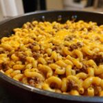 Close-up of Cheesy Ground Beef Pasta Skillet Comfort Food featuring elbow macaroni, ground beef, and melted cheddar cheese in a black skillet.