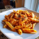 A serving of vibrant orange Creamy Cajun Pasta with Chicken and Spice, topped with dried herbs, on a white square plate.