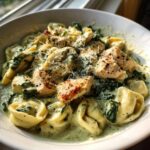 A close-up of a white bowl filled with Creamy Chicken Tortellini with Spinach Sauce, topped with grilled chicken chunks and grated Parmesan.