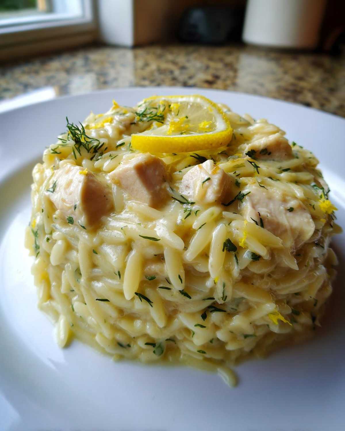 A serving of Creamy Lemon Chicken Orzo, molded and topped with chicken pieces, dill, and a lemon slice.