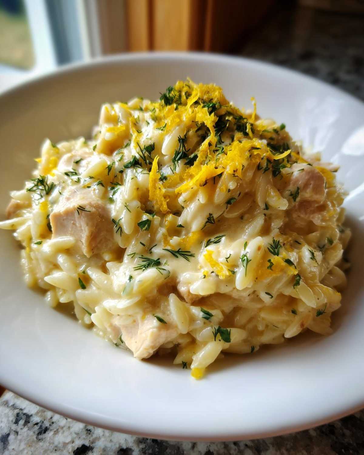 Close-up of a bowl of Creamy Lemon Chicken Orzo topped with bright yellow lemon zest and fresh green herbs.