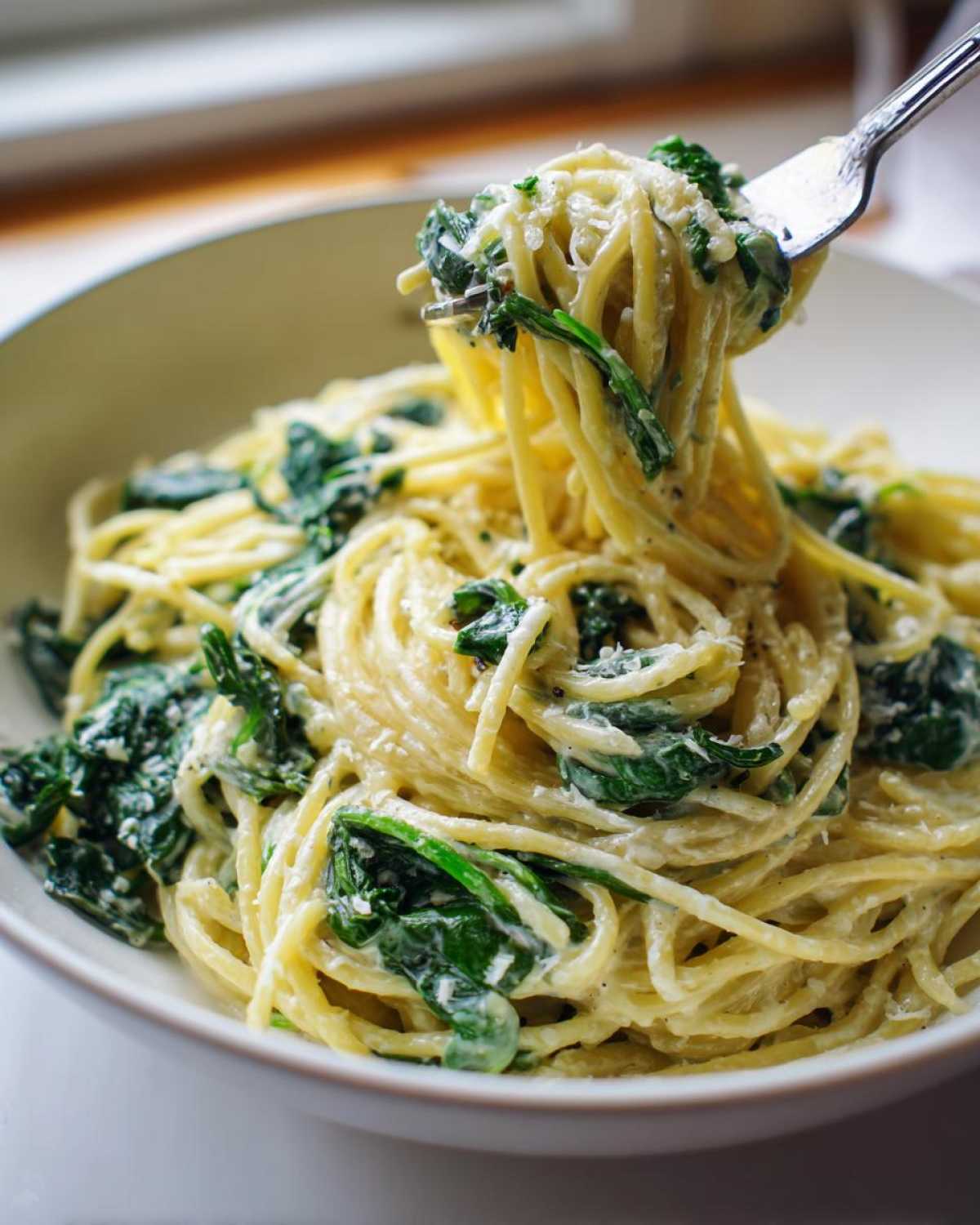 A fork lifting a twirl of spaghetti coated in creamy sauce, spinach, and cheese from a bowl of Creamy Lemon Ricotta Pasta with Spinach.
