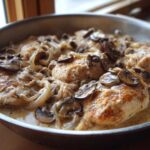 Close-up of seared chicken breasts covered in a rich, creamy mushroom sauce with sliced onions in a skillet.