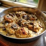 Close-up of seared chicken breasts covered in a rich, creamy mushroom sauce in a stainless steel skillet.