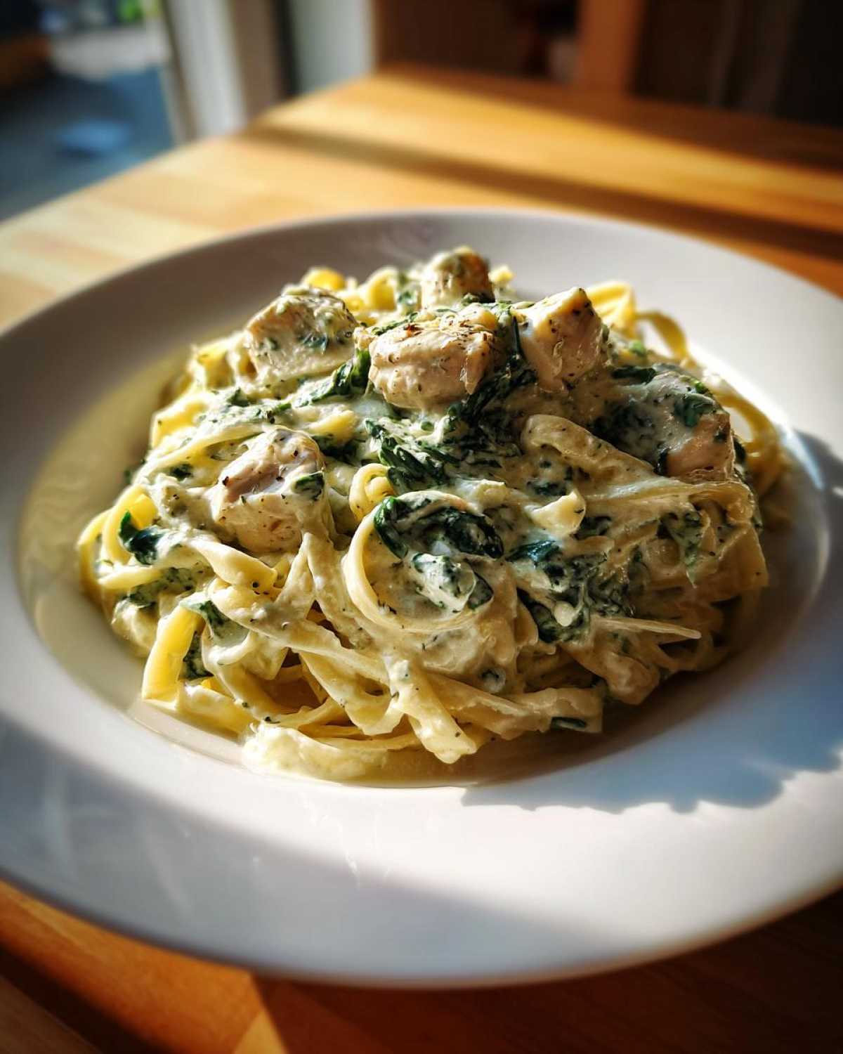 A close-up of a white bowl filled with Creamy Spinach Chicken Pasta with Garlic, featuring fettuccine, chunks of chicken, and spinach in a rich white sauce.