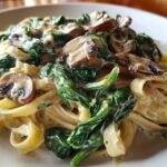 A close-up of fettuccine coated in a rich white sauce, topped with wilted spinach and sliced mushrooms, ready for a Creamy Spinach and Mushroom Pasta Dinner.