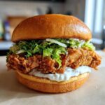 A close-up of a delicious Crispy Chicken Burger with Garlic Mayo, featuring a thick fried patty, shredded lettuce, and a brioche bun.