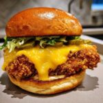 A close-up of a delicious Crispy Chicken Burger with Melty Cheese, lettuce, and pickles on a brioche bun.