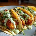 Three Crispy Chicken Tacos topped with creamy avocado sauce and fresh cilantro on a gray plate.