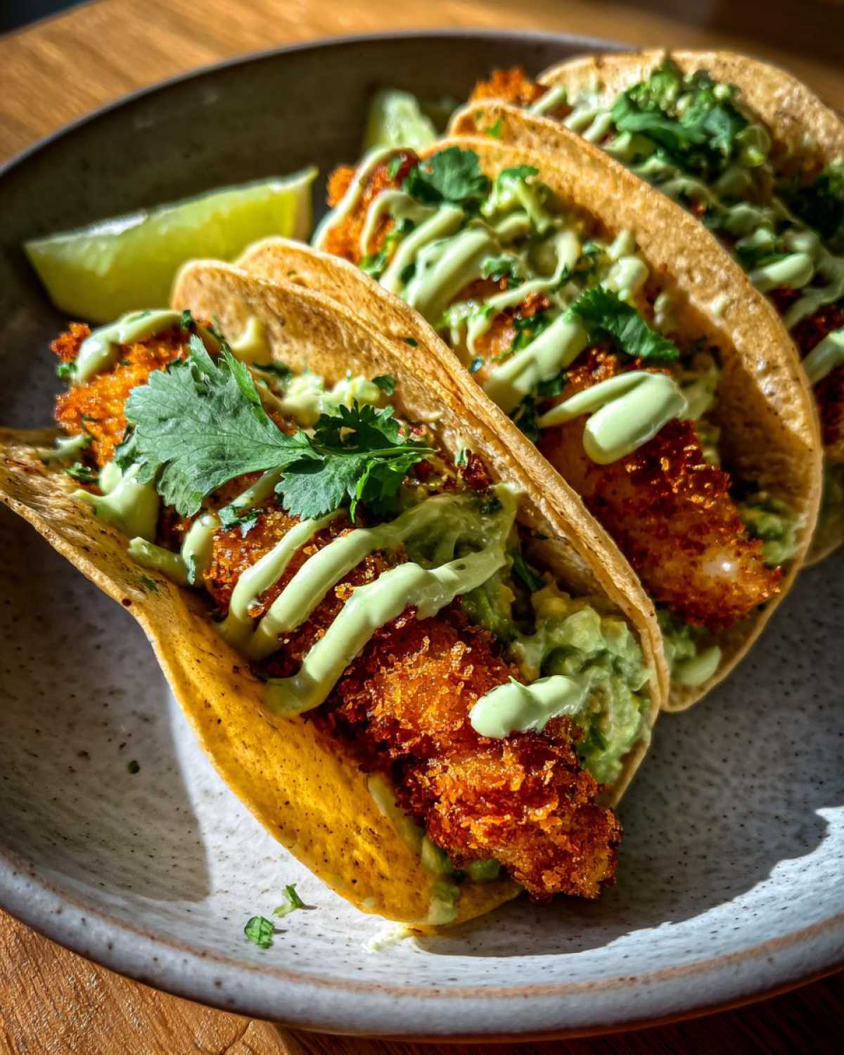 Three Crispy Chicken Tacos with avocado cream drizzle and cilantro on a speckled plate, served with a lime wedge.
