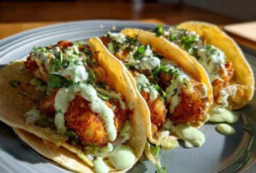 Three Crispy Chicken Tacos topped with creamy avocado sauce and fresh cilantro on a gray plate.