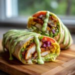 Two halves of a Crispy Chicken Wrap with Creamy Avocado Sauce drizzled generously over the top, resting on a wooden board.