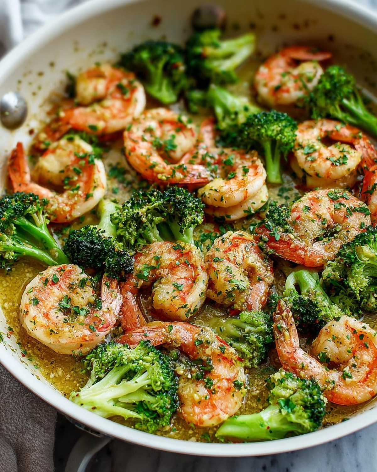 Close-up of plump shrimp and bright green broccoli florets simmering in a rich garlic butter sauce in a white skillet.