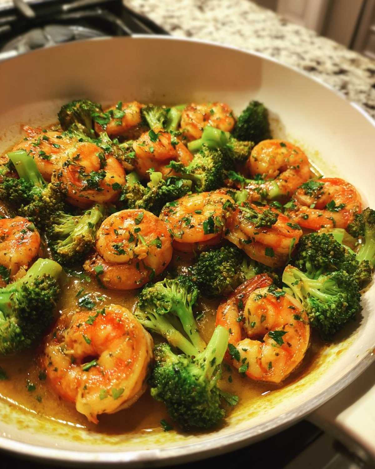 Close-up of cooked shrimp and bright green broccoli florets coated in garlic butter sauce in a white skillet.