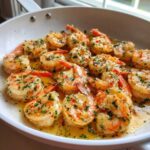 Close-up of cooked shrimp coated in garlic butter sauce, herbs, and red pepper flakes in a white skillet.
