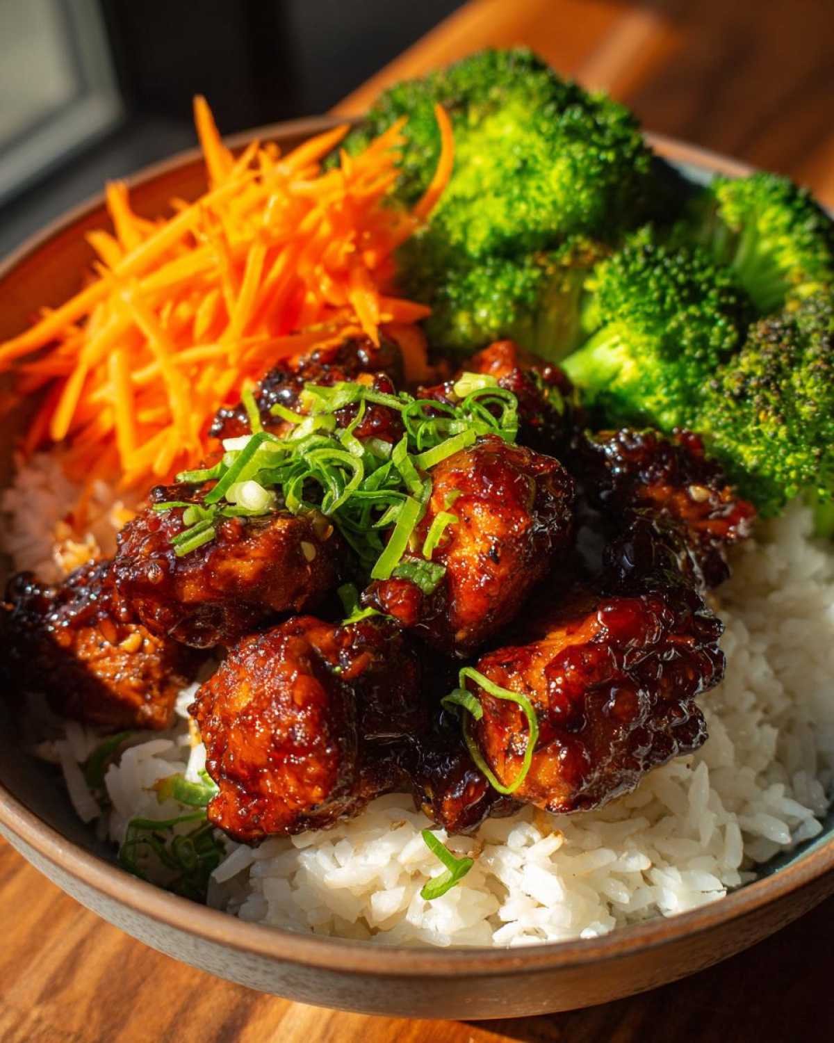 Close-up of a Honey Garlic Chicken Bowl featuring sticky glazed chicken pieces over white rice, served with bright green broccoli and shredded carrots.