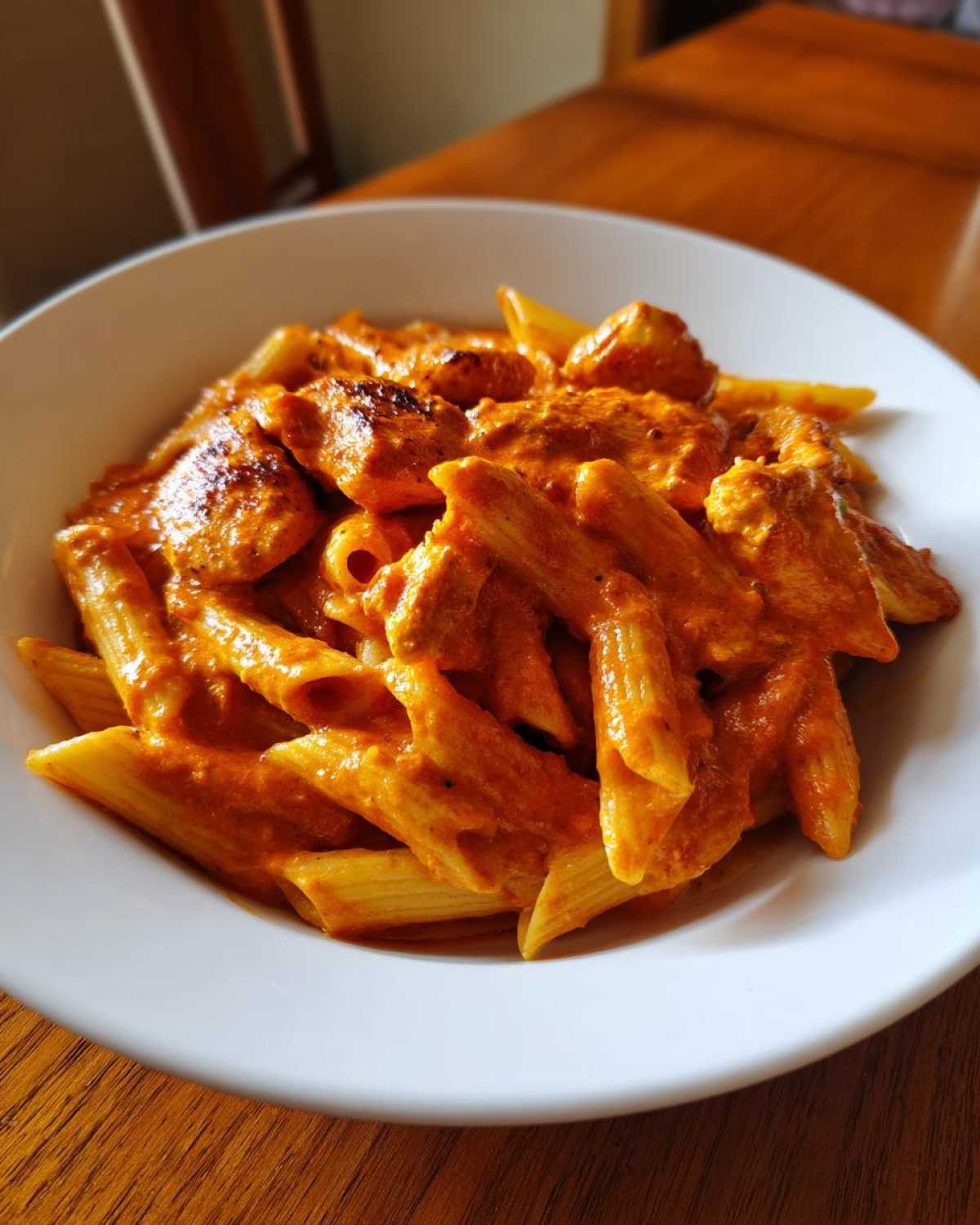 Close-up of penne pasta coated in rich, creamy tomato sauce with chunks of chicken, served in a white bowl.