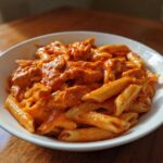 A close-up of a white bowl filled with One Pot Creamy Tomato Pasta with Chicken, featuring penne pasta and chunks of chicken in a rich orange sauce.