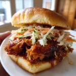 A close-up of a Sticky BBQ Chicken Sandwich piled high with saucy chicken and crunchy coleslaw on a brioche bun.