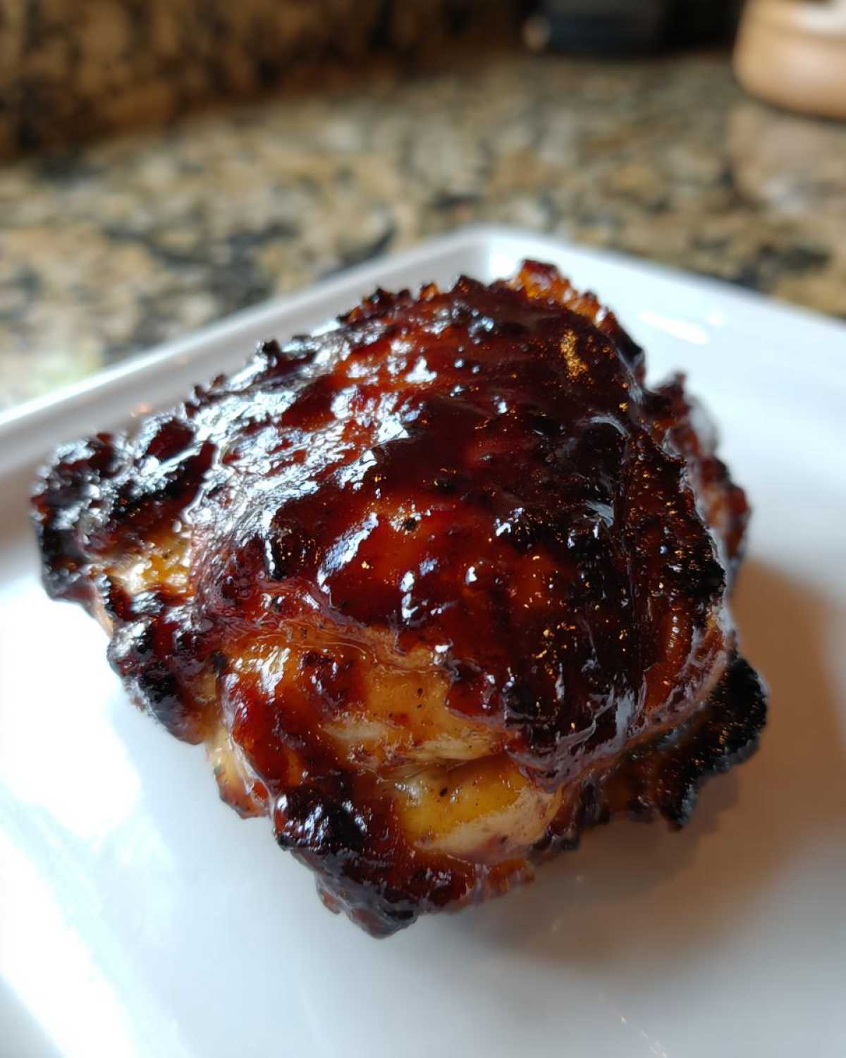 A single, glistening Sticky BBQ Chicken Thighs piece with a dark, caramelized, and crispy BBQ glaze on a white plate.