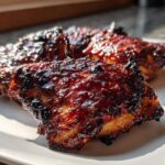 Three pieces of Sticky BBQ Chicken Thighs with crispy, caramelized edges glistening under bright sunlight on a white plate.