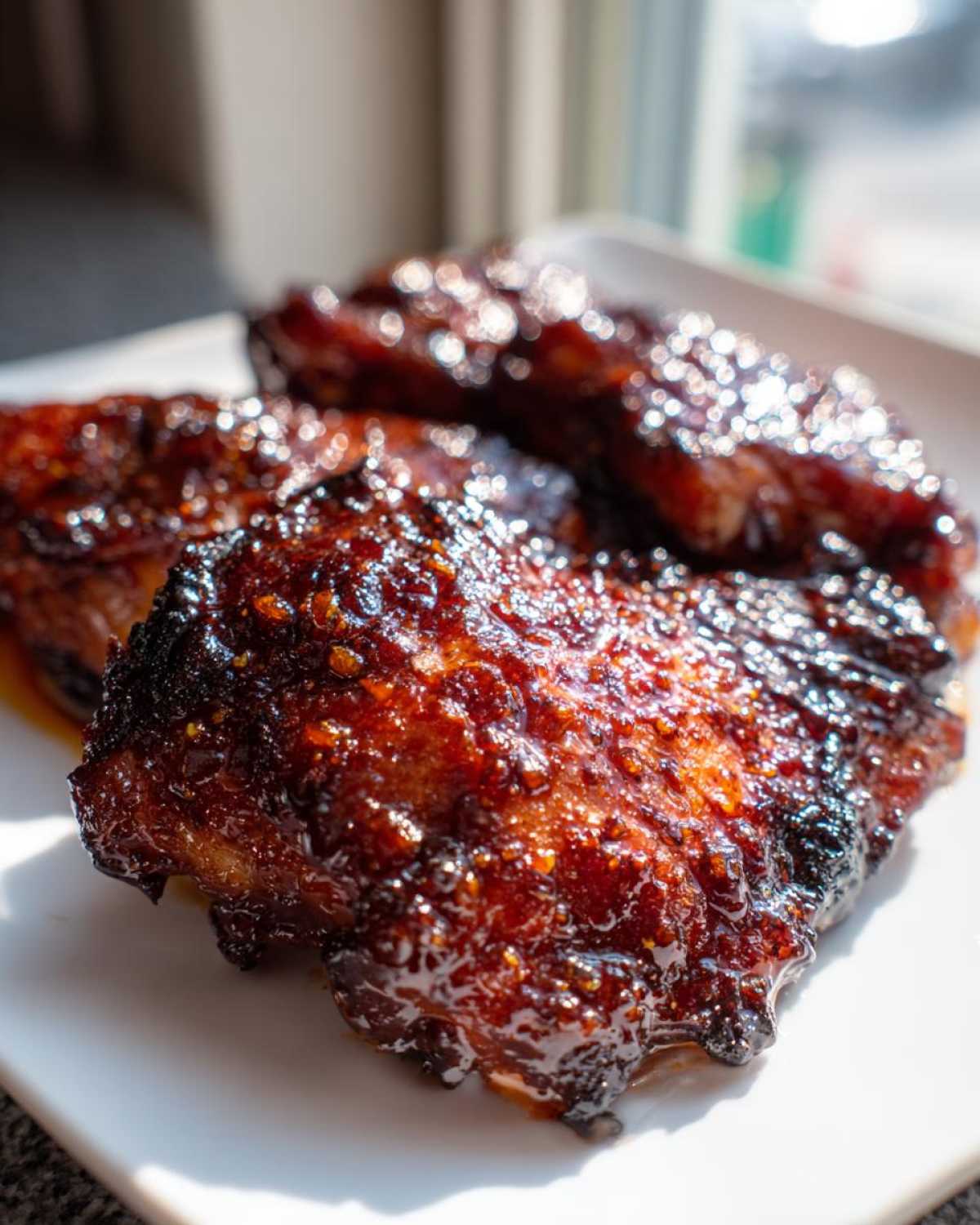 Close-up of glistening, dark brown Sticky BBQ Chicken Thighs with crispy, caramelized edges on a white plate.
