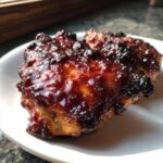 Two pieces of Sticky BBQ Chicken Thighs with Crispy Edges glistening under bright light on a white plate.