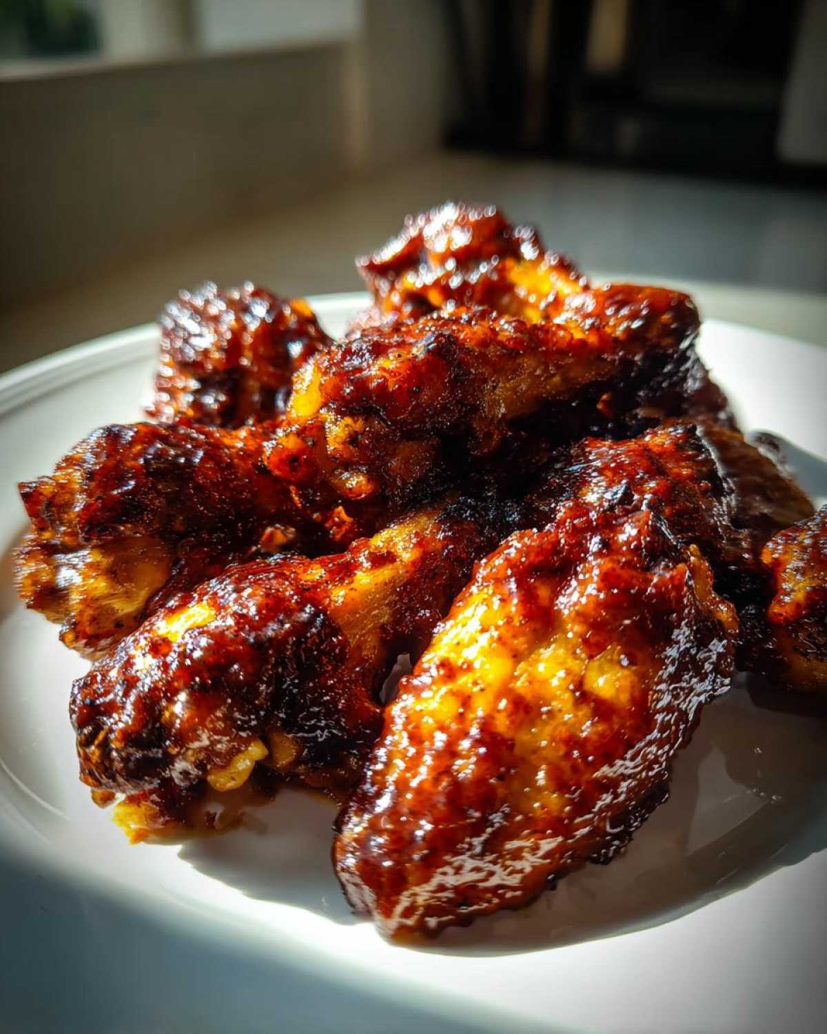 A close-up of several Sticky BBQ Chicken Wings Crispy Oven Style coated in a dark, glossy sauce on a white plate.