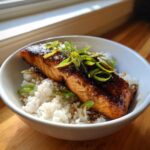 A close-up of a perfectly cooked Sticky Honey Soy Salmon fillet served over white rice and garnished with fresh green onions.