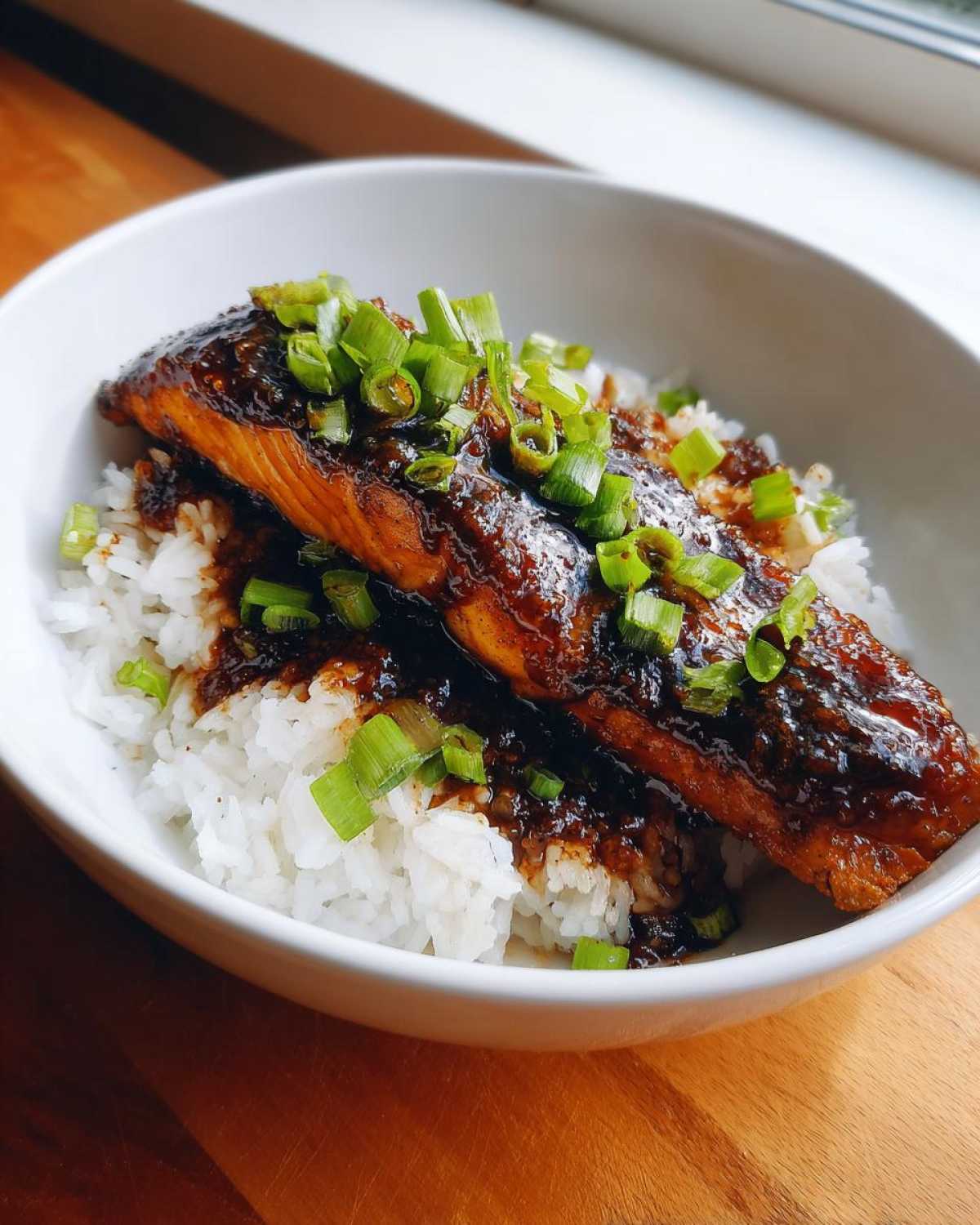 A close-up of a Sticky Honey Soy Salmon fillet glazed with dark sauce, served over white rice and topped with fresh green onions.