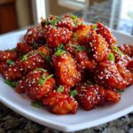 A mound of Sticky Sweet Chili Chicken Easy Recipe pieces coated in glossy red sauce, topped with sesame seeds and green onions.
