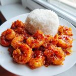 A plate featuring glistening Sticky Sweet Chili Shrimp coated in a vibrant red sauce next to a mound of white rice.
