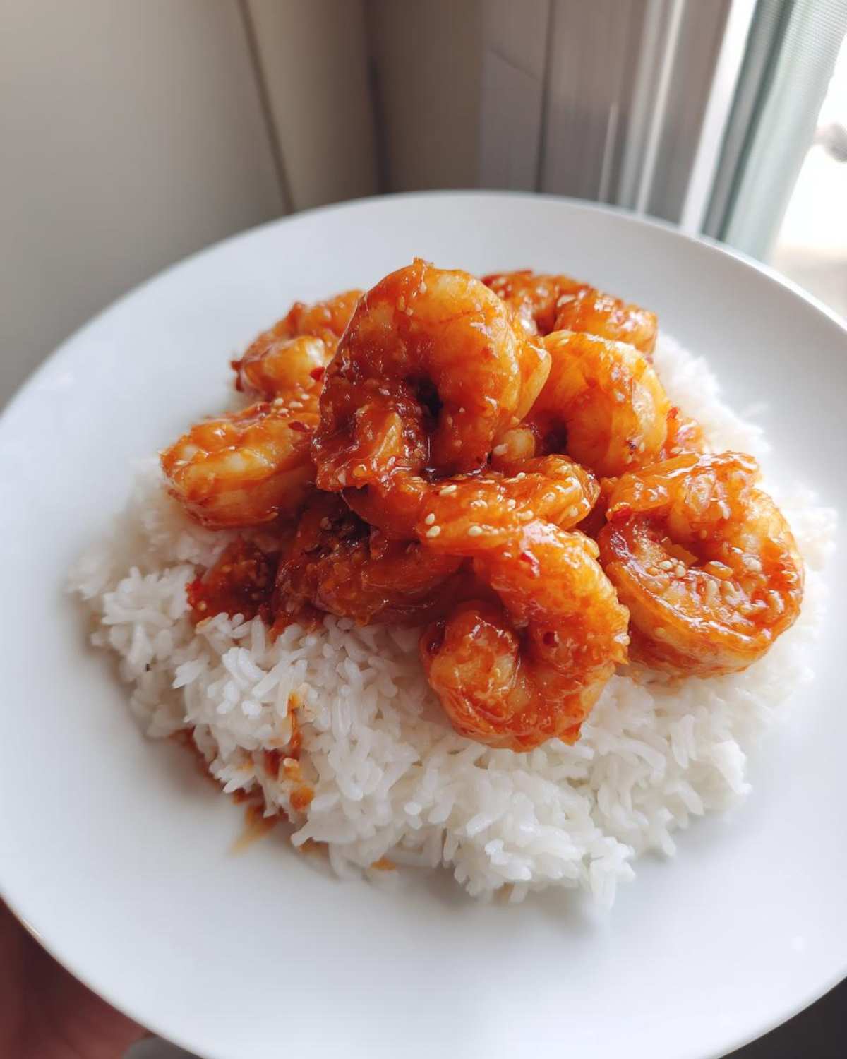 Close-up of Sticky Sweet Chili Shrimp coated in glaze and sesame seeds, served over a bed of fluffy white rice on a white plate.