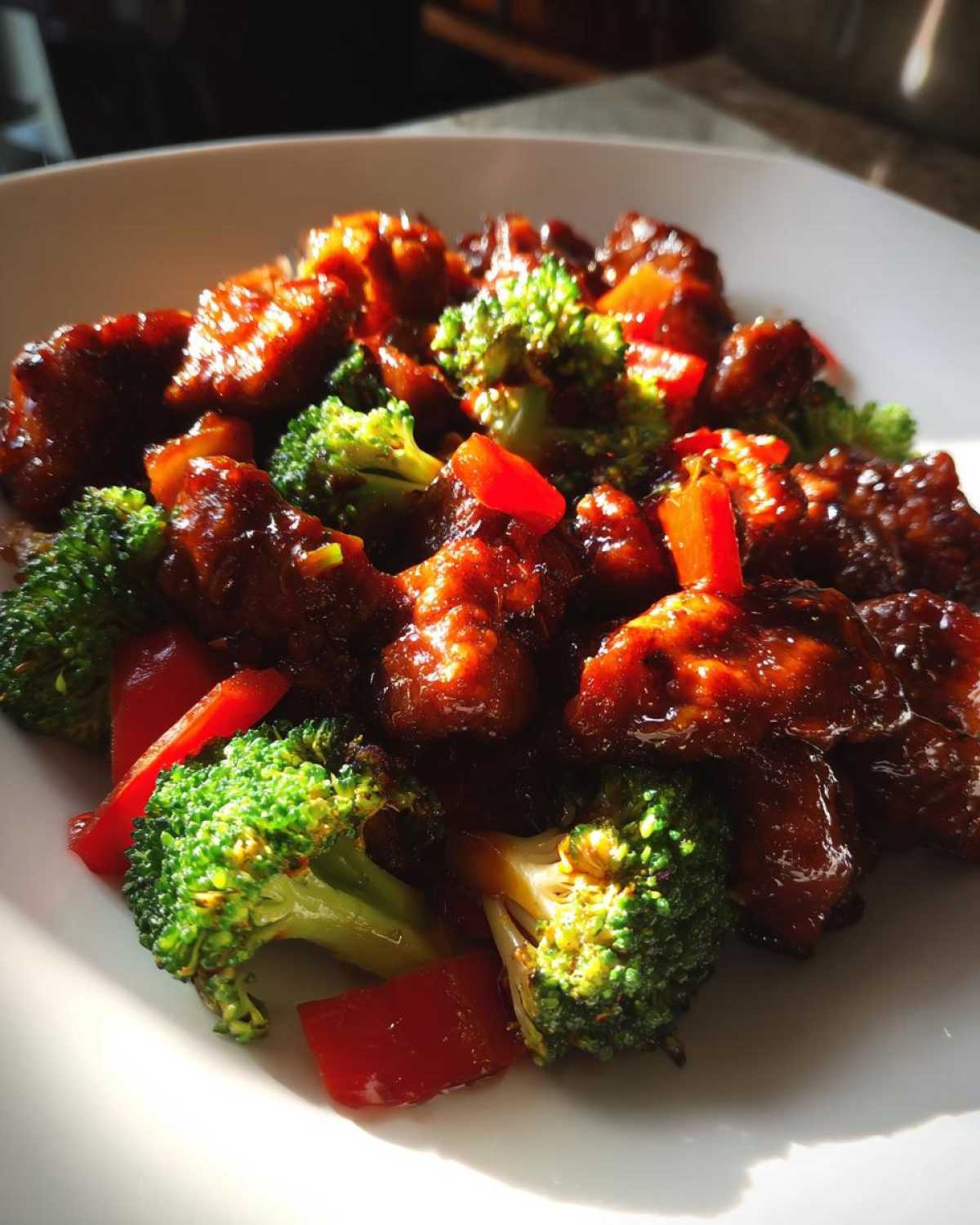 Close-up of glistening Sticky Teriyaki Chicken Stir Fry mixed with bright green broccoli florets and red pepper chunks on a white plate.