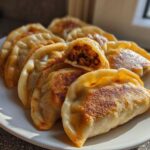 A plate of golden brown, pan-fried dumplings, with one dumpling cut open showing the savory filling.